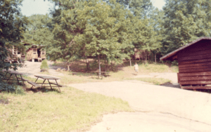 Wooden cabins, picnic tables, wooded area.