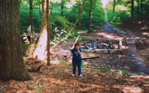 Woman in woods near campfire remains.