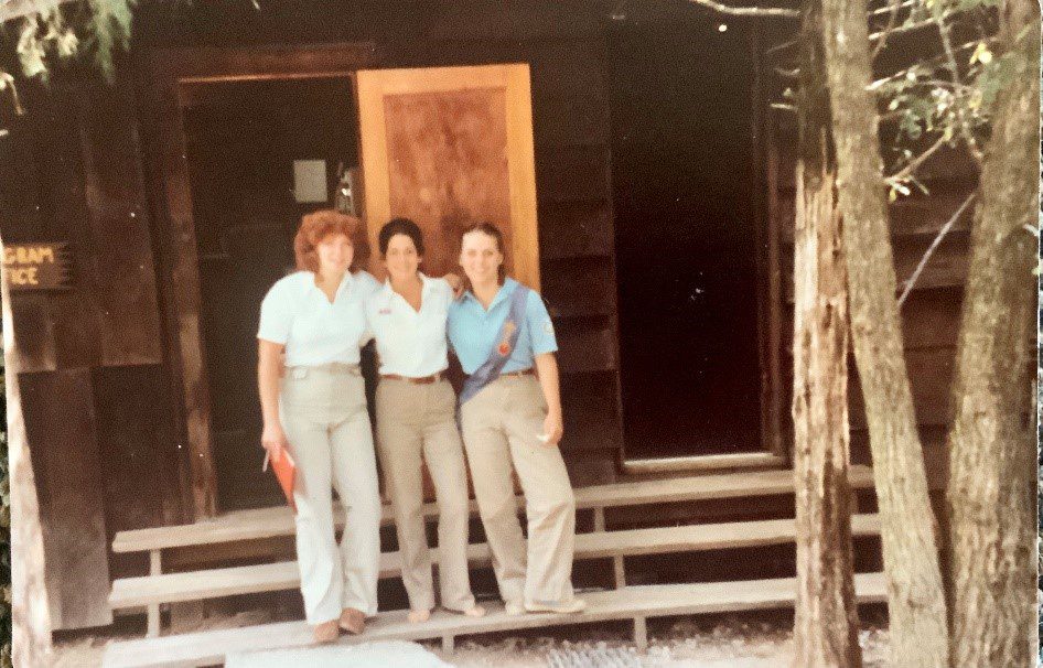 Three women stand outside a cabin.