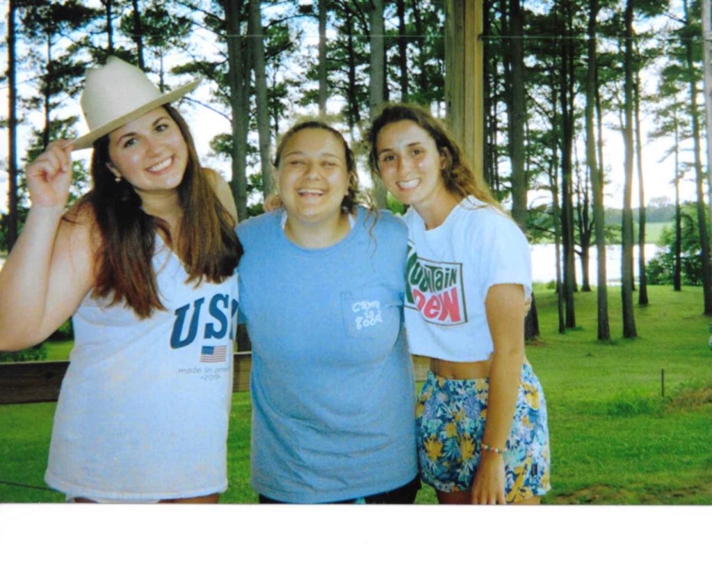 Three friends smiling by lakeside among pine trees