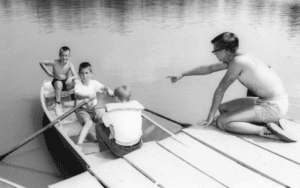 Boys rowing a boat, man watching.