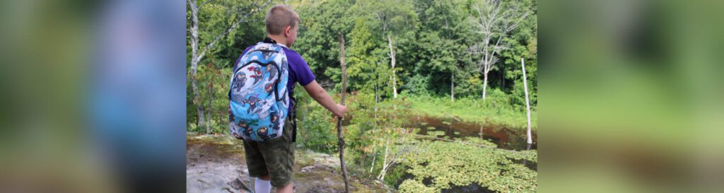 Young boy hikes with backpack and stick.