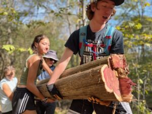Teenagers carrying large logs in woods.