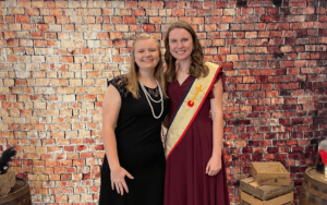Two women in formal wear, brick wall background.